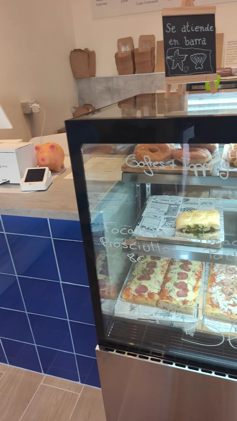 Mostrador con azulejos azules y vitrina de focaccias, donuts y gofres, junto a la pizarra «Se atiende en barra»